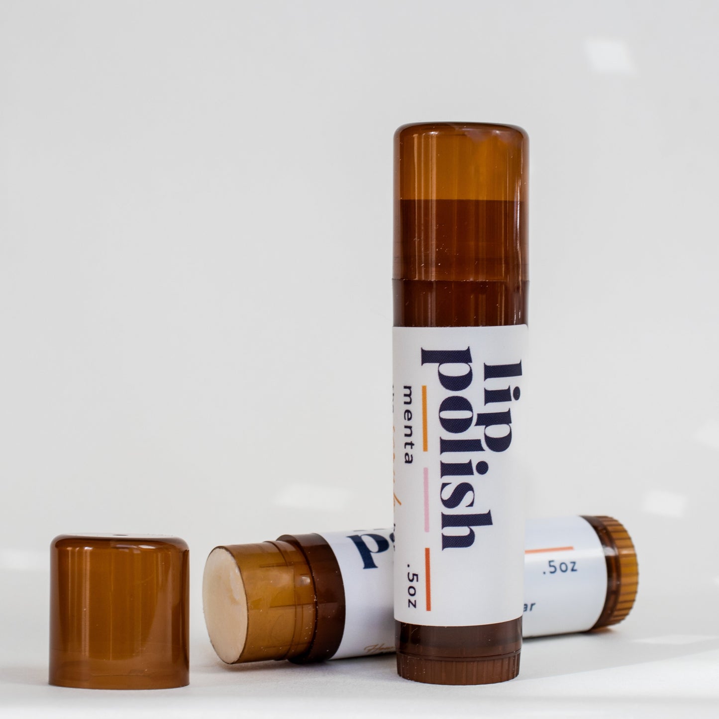 A close-up image of The Scrub Bar's Lip Polish in a .5oz amber tube, showcasing the (menta) peppermint-scented lip balm with the cap removed. The tube sits against a clean, white background with soft light reflections, highlighting the smooth and glossy texture of the product.

