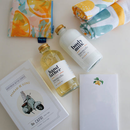 The Scrub Bar Featuring- The Capri Tasting Gift Set featuring lemon-patterned kitchen towels, Capri Lemon Poppy Cookies, lemon-themed stationery notepad, Limoncello Hand Lather, Limoncello Body Milk, and a reusable lemon-patterned bag. A perfect lemon kitchen theme gift basket from The Scrub Bar.