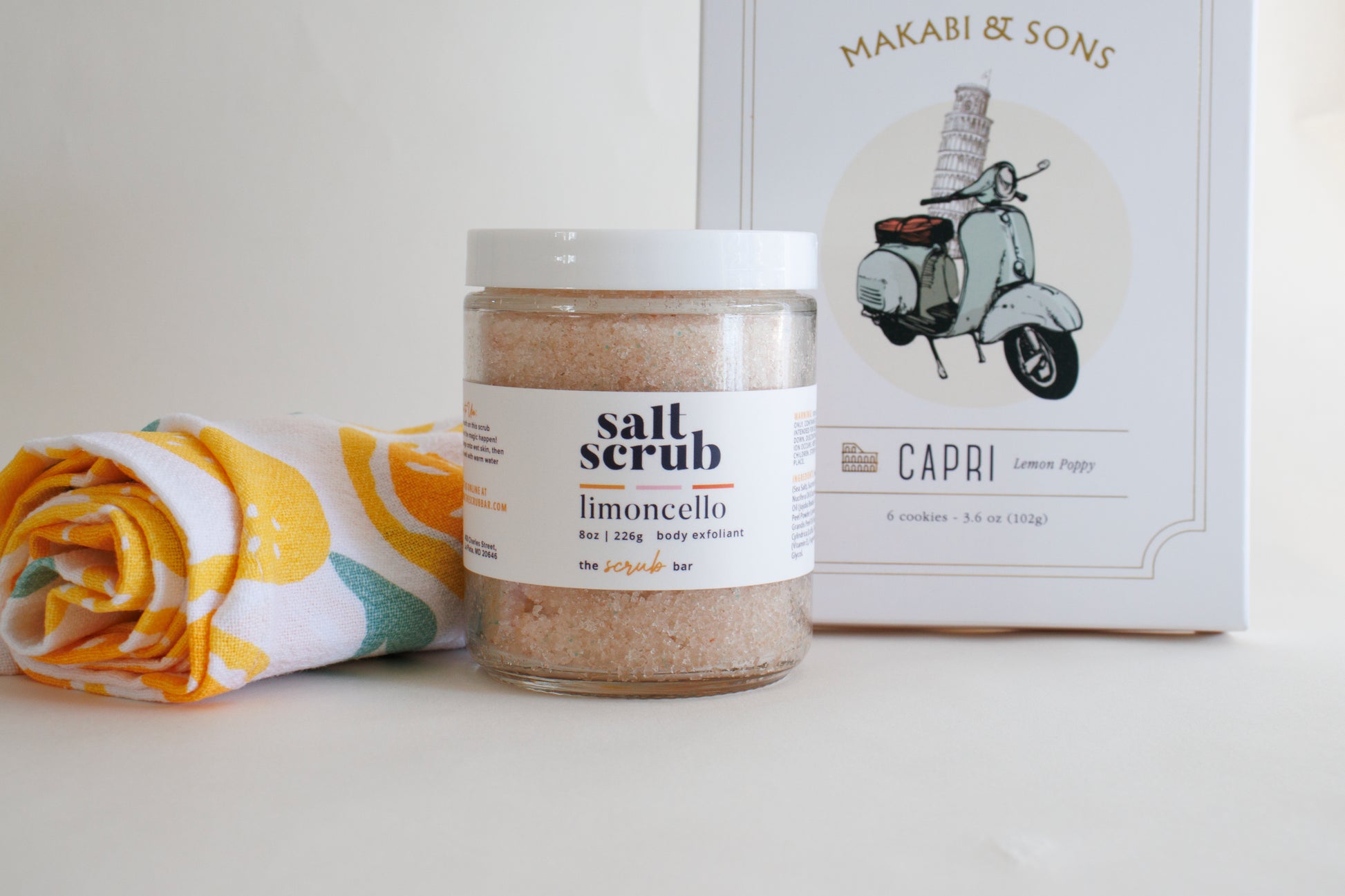 The Scrub Bar's Lemon Verbena Gift Set featuring 1 assorted lemon-patterned kitchen towel, 8oz Limoncello exfoliating salt scrub, and Capri Lemon Poppy Cookies. The perfect hostess gift, gifts under $50, or gifts for mature women