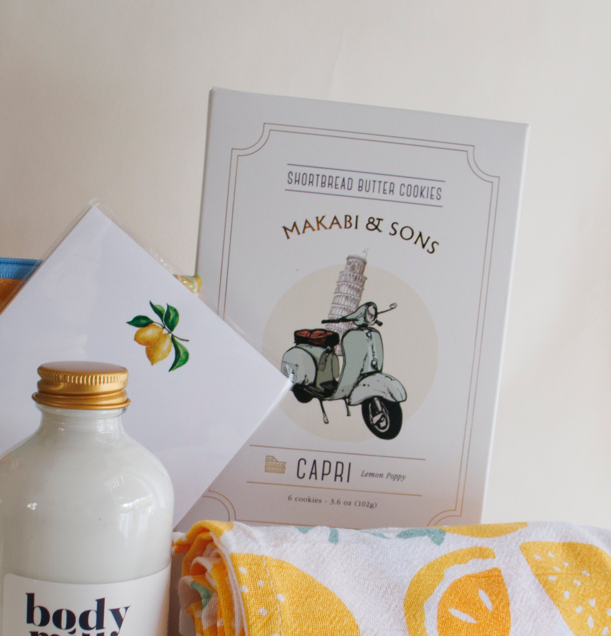 The Scrub Bar Featuring- Makabi & Sons Capri Lemon Poppy Cookies, Limoncello Hand Lather, and Limoncello Body Milk arranged as part of The Capri Tasting Gift Set from The Scrub Bar. This lemon kitchen theme gift basket includes lemon-patterned kitchen towels, stationery, and a reusable lemon-patterned bag.